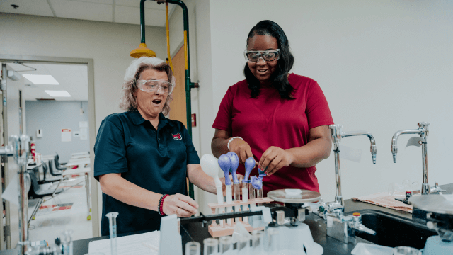 a teacher wearing goggles and a red shirt standing next to a student wearing goggles